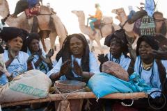 Des femmes font du tendé tandis que les hommes paradent autours d'elles sur des dromadaires, festival de l'Aïr, Iférouane, 2019.